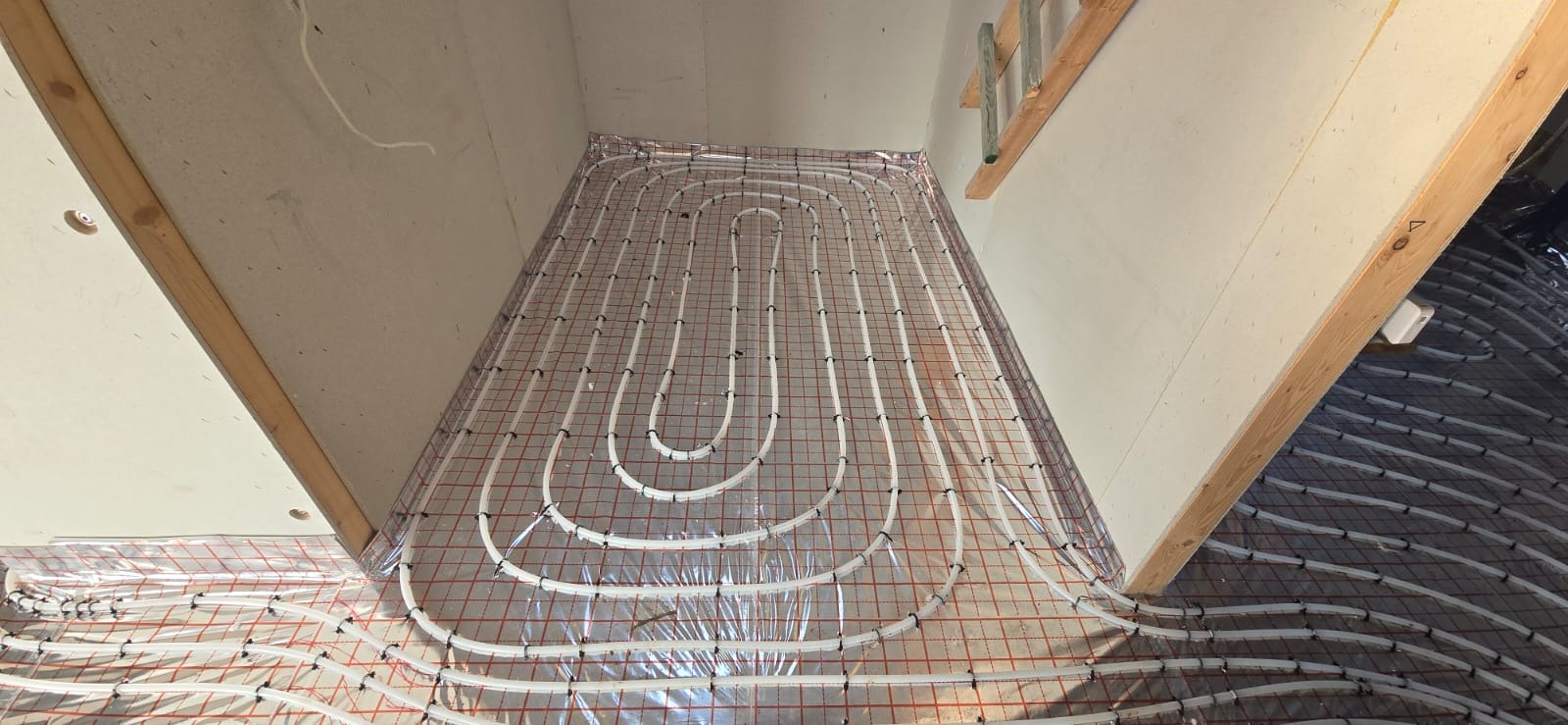 Białe rury ogrzewania podłogowego ułożone na folii z czerwoną siatką, widoczne między drewnianymi elementami konstrukcyjnymi. Część instalacji poza kadrem.