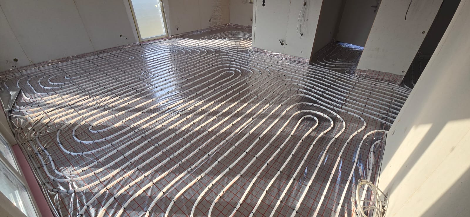 Ułożona instalacja ogrzewania podłogowego z białych rurek na czerwonej siatce, widok z góry, surowy stan wykończenia wnętrza, przygotowanie do wylewki.