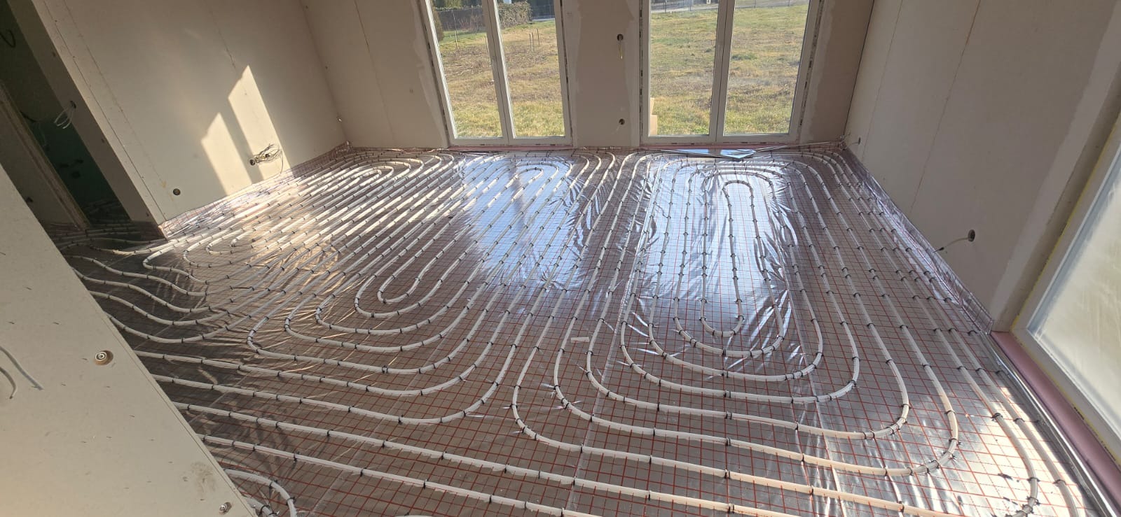 Białe rury ogrzewania podłogowego ułożone na folii z czerwoną siatką w jasnym pokoju z oknami. Widoczne ściany w stanie surowym i wystające kable.