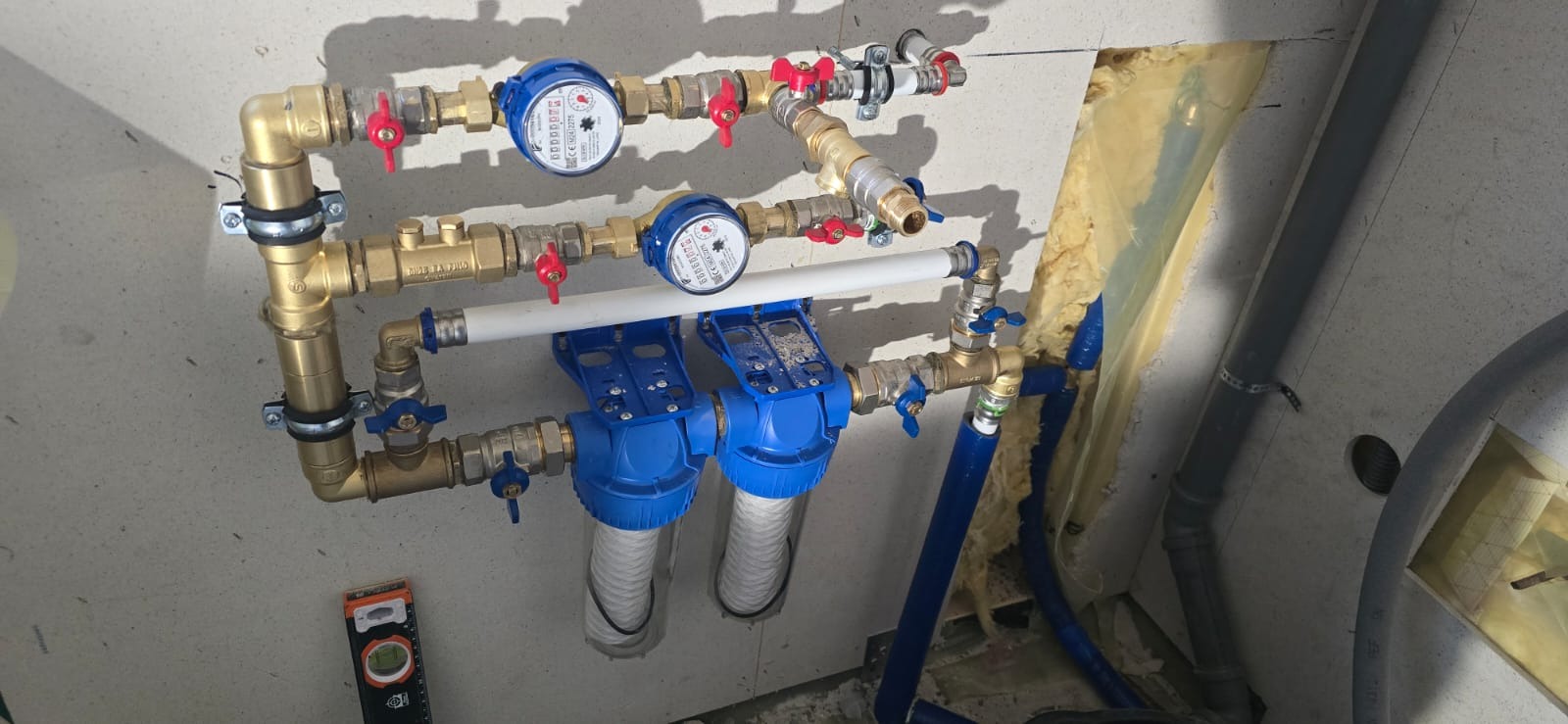 Kompletna instalacja wodna z filtrami i licznikami, mosiężne rury, czerwone i niebieskie zawory, poziomica przy ścianie. Widoczne elementy instalacji w surowym stanie.