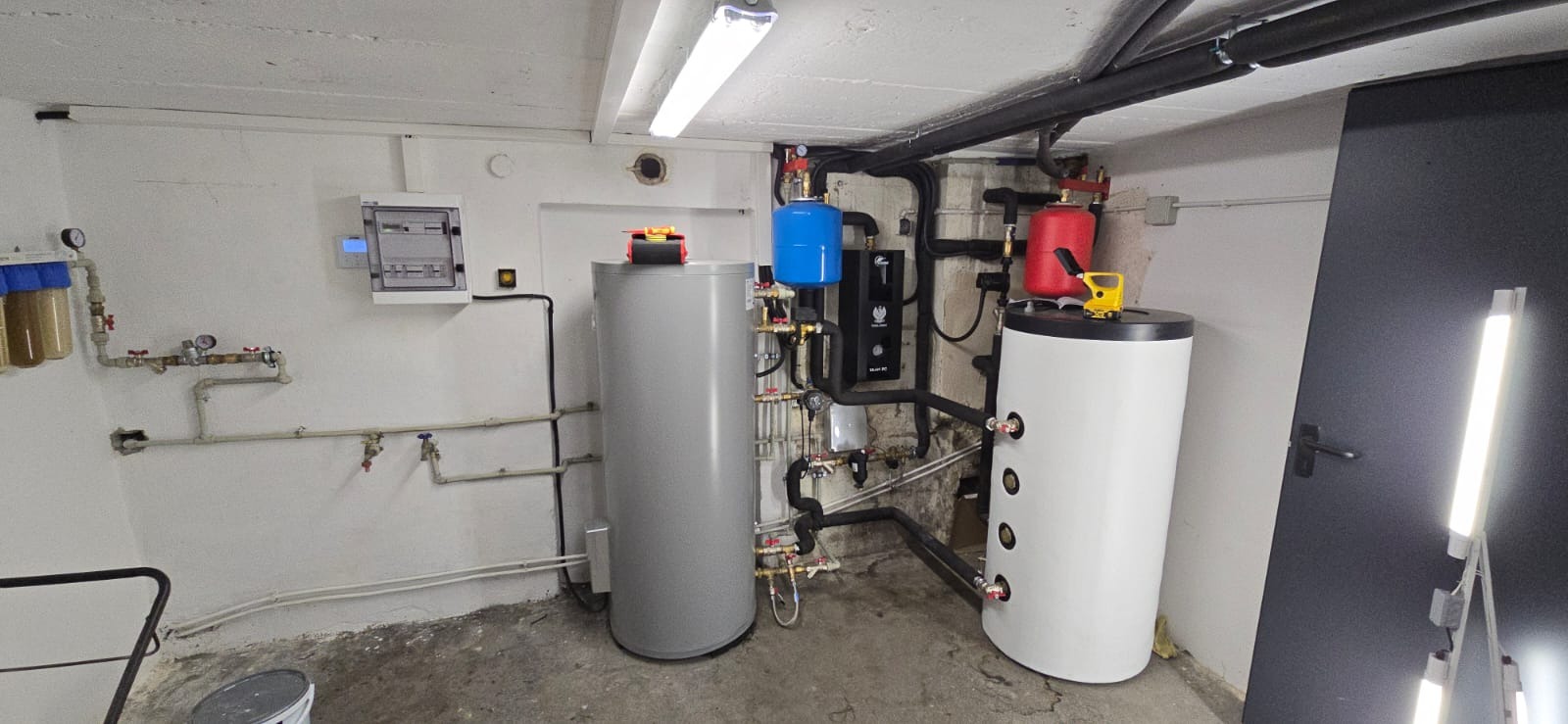 Nowoczesna kotłownia z dwoma zbiornikami, orurowaniem, filtrami i rozdzielnicą elektryczną na ścianie. Widoczne elementy instalacji grzewczej i sanitarnej w piwnicy.