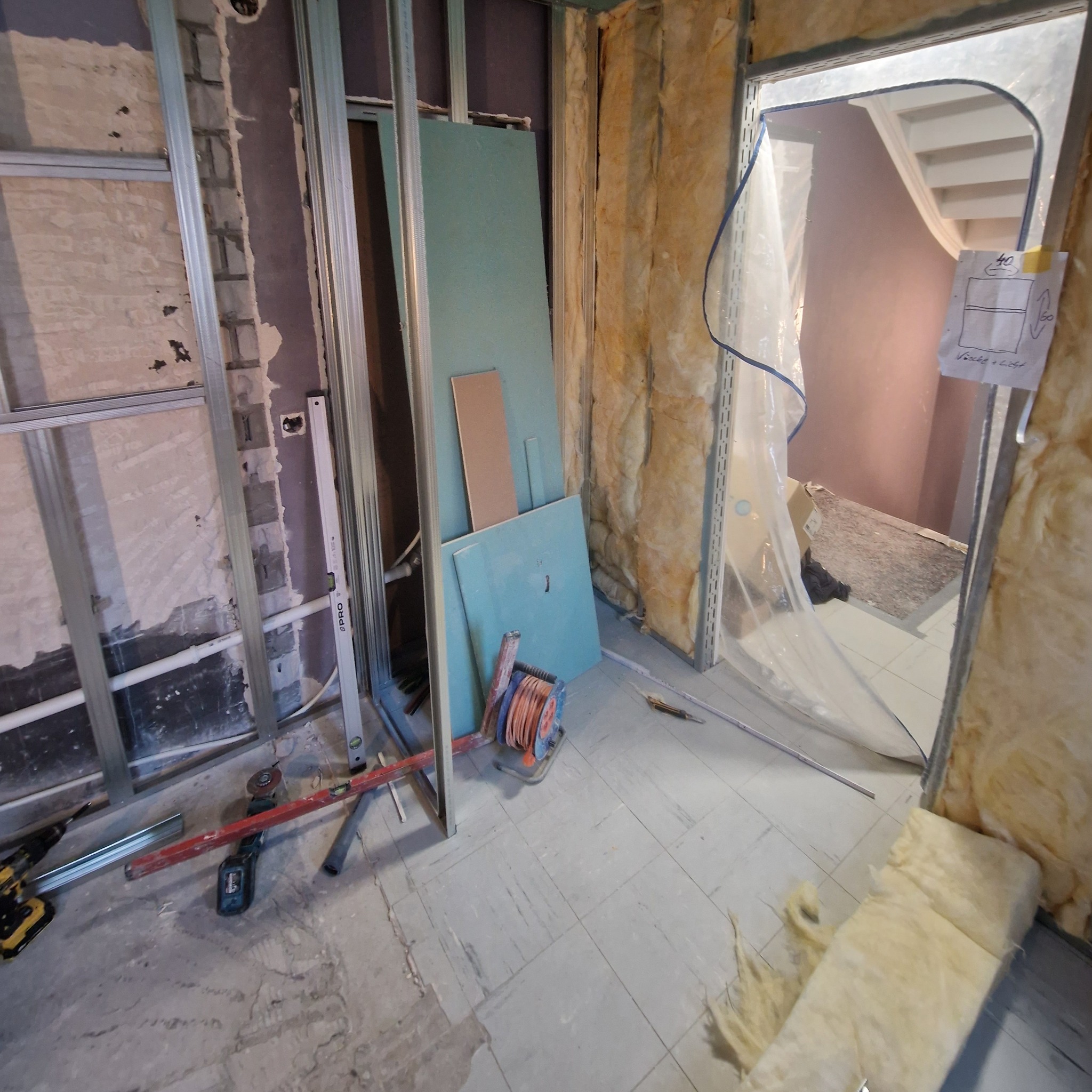 Wnętrze w trakcie prac wykończeniowych: izolacja ścian wełną mineralną, stelaż pod płyty gipsowe, narzędzia i materiały budowlane na podłodze. Widok na schody przez otwór w ścianie.