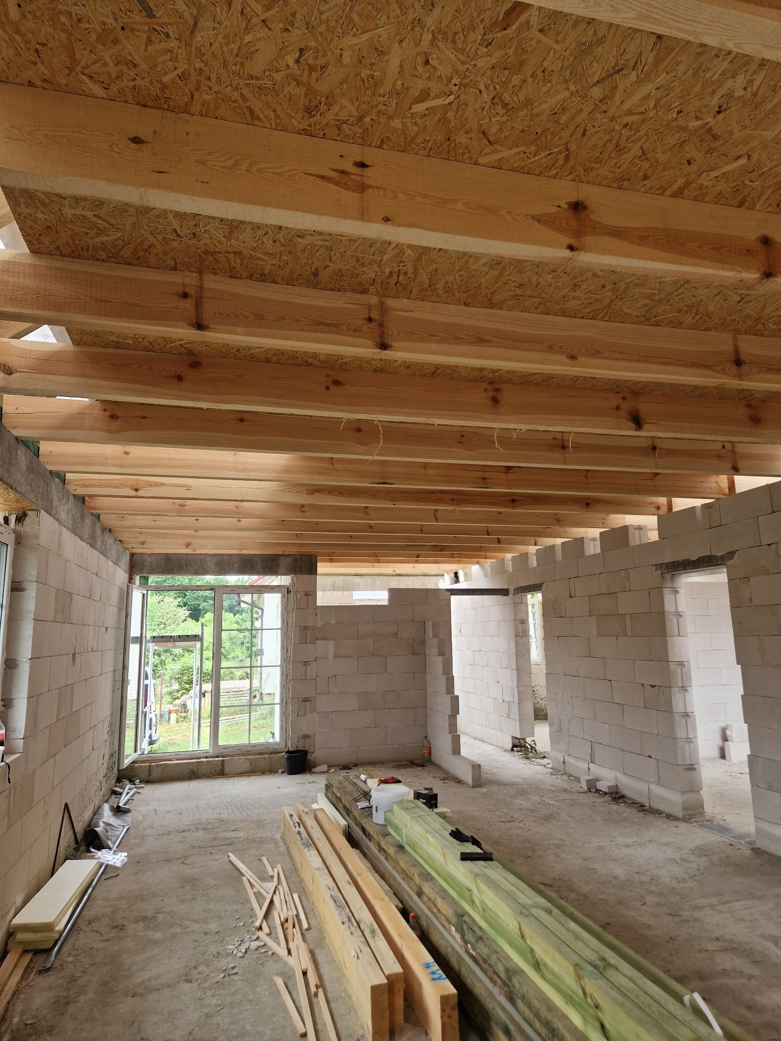 Wnętrze surowego domu w trakcie budowy: ściany z betonu komórkowego, drewniany strop z desek OSB, okna z widokiem na zieleń. Na podłodze leżą materiały budowlane.