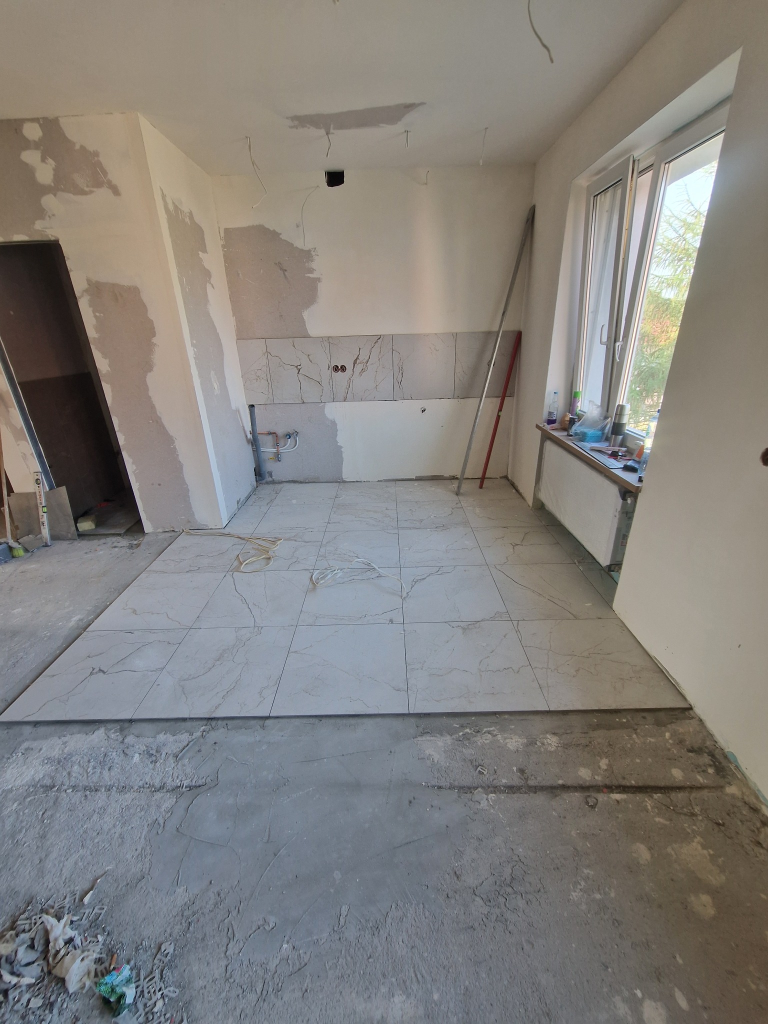 Wnętrze w trakcie remontu: ułożone płytki podłogowe imitujące marmur, częściowo wykończona ściana z płytkami nad blatem kuchennym, surowe ściany i podłoga.