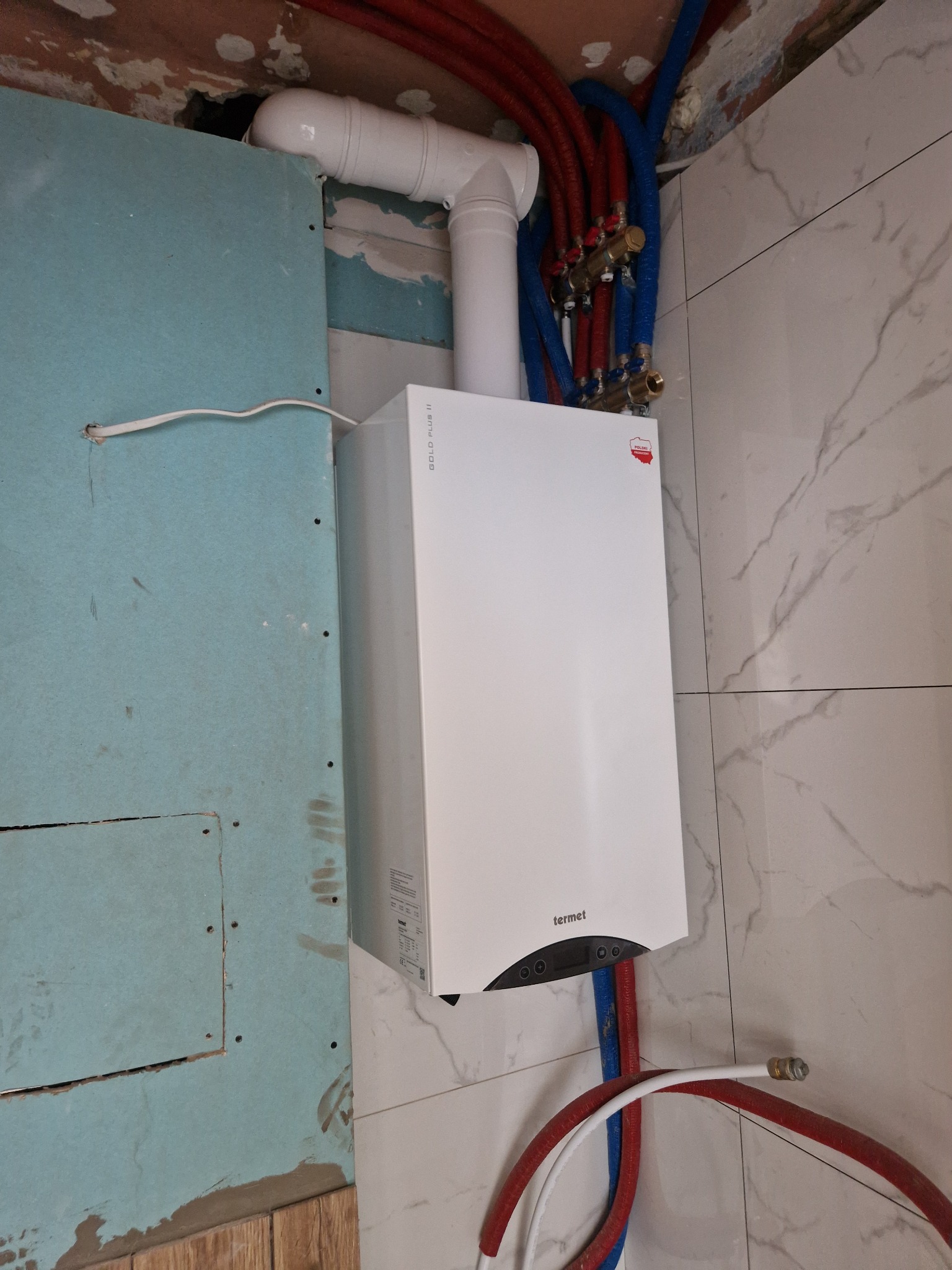Biały piec gazowy Termet na tle marmurowych płytek i niebieskiej płyty karton-gips z widocznymi instalacjami hydraulicznymi w kolorze czerwonym i niebieskim.