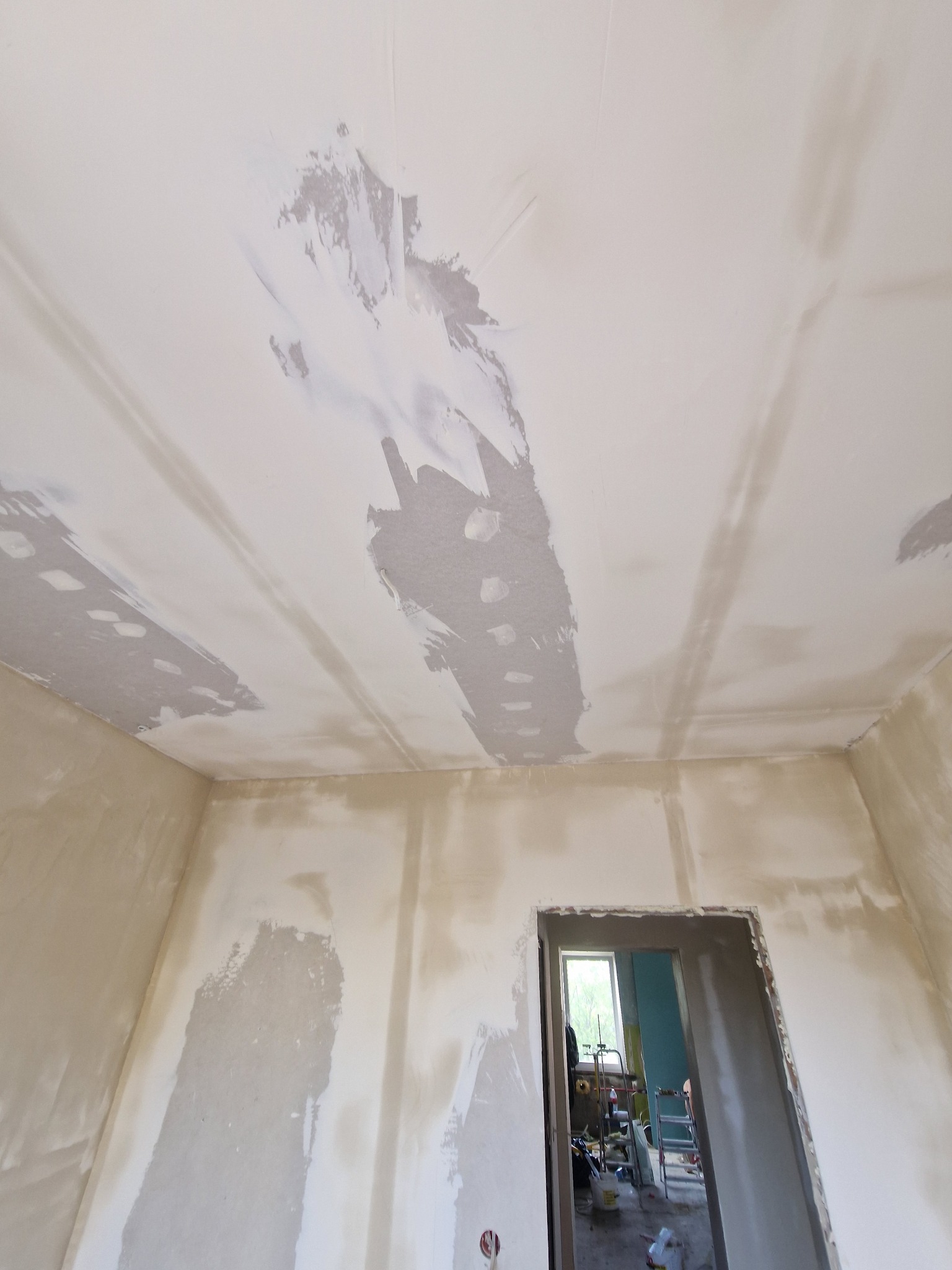 Wnętrze w trakcie szpachlowania: sufit z widocznymi łączeniami płyt gipsowych, ściany w jasnej szpachli, widok na drzwi do innego pomieszczenia. Prace wykończeniowe.