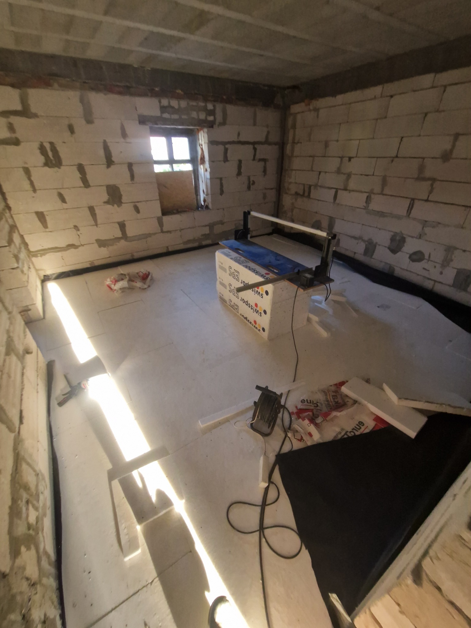 Ocieplenie podłogi styropianem w surowym wnętrzu. Widoczna maszyna do cięcia styropianu, folia izolacyjna i fragment okna. Ściany z pustaków.