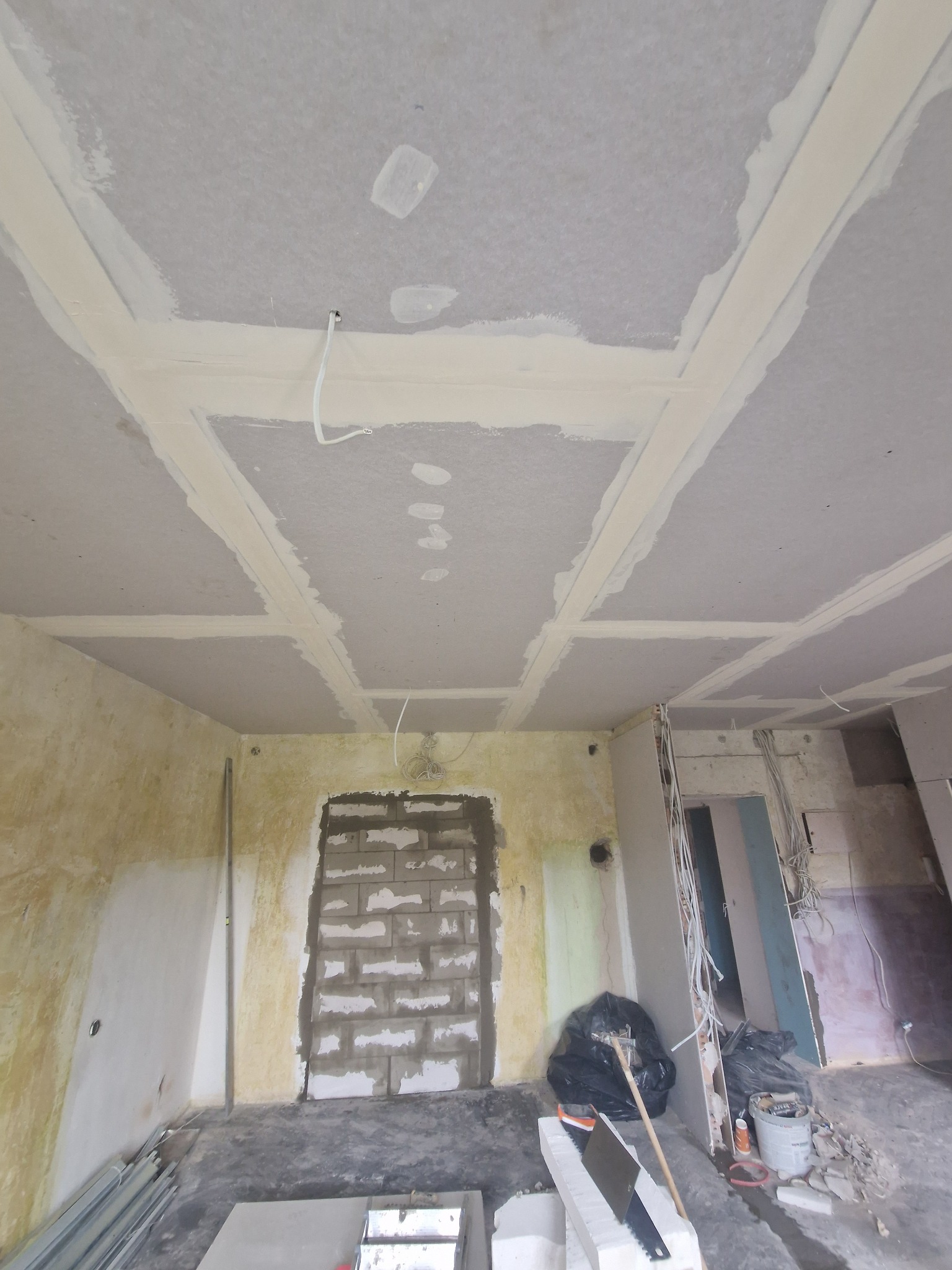 Sufit z płyt kartonowo-gipsowych w trakcie montażu, widoczne łączenia i przewody elektryczne. Ściany w surowym stanie, narzędzia i materiały budowlane w pomieszczeniu.