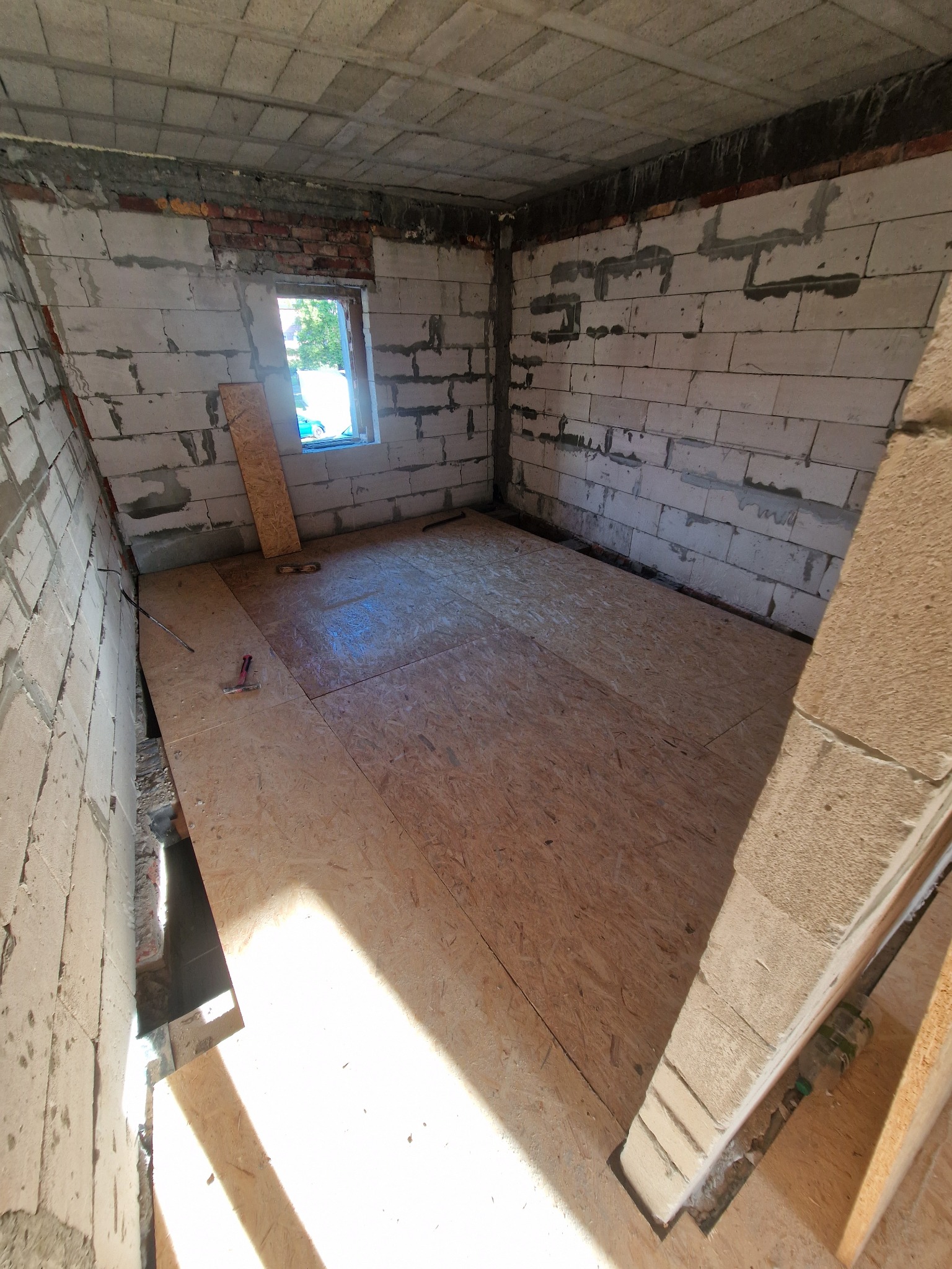 Surowy stan poddasza z oknem i częściowo ułożoną podłogą z płyt OSB. Widoczne narzędzia i niedokończone ściany z betonu komórkowego.