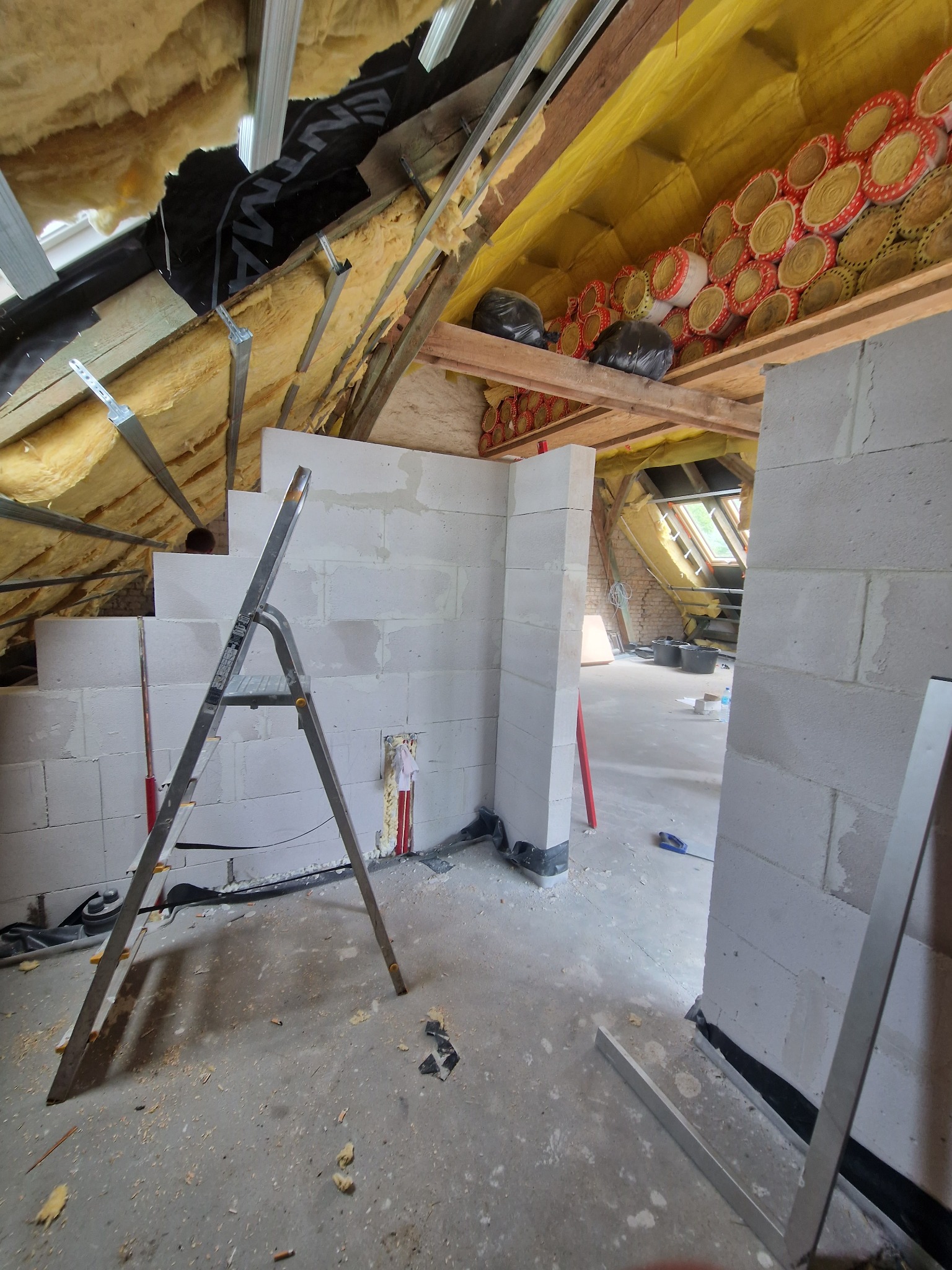 Adaptacja poddasza: ściany z bloczków, ocieplenie wełną mineralną, drabina. Widoczne elementy konstrukcji dachu i okno dachowe. Prace w toku, surowy stan.