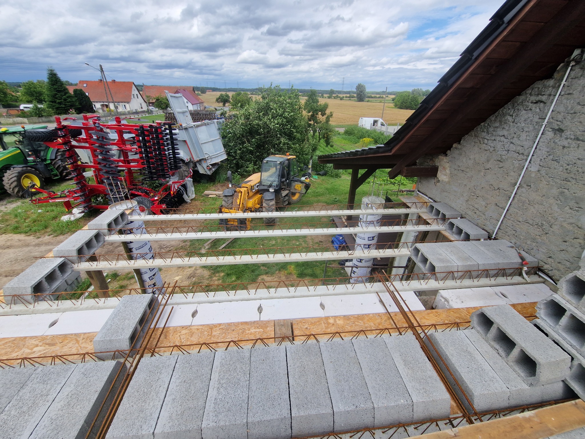 Budowa stropu Teriva z pustaków szalunkowych, zbrojenia i izolacji. W tle maszyny rolnicze i budynki mieszkalne. Widok z góry na plac budowy w słoneczny dzień.
