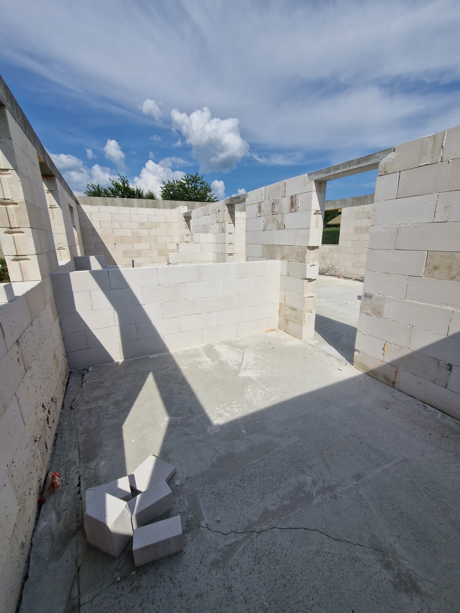 Wnętrze surowego domu w trakcie budowy. Ściany z białych bloków, betonowa podłoga z pęknięciami, fragmenty bloczków na podłodze. Widok na błękitne niebo z chmurami.