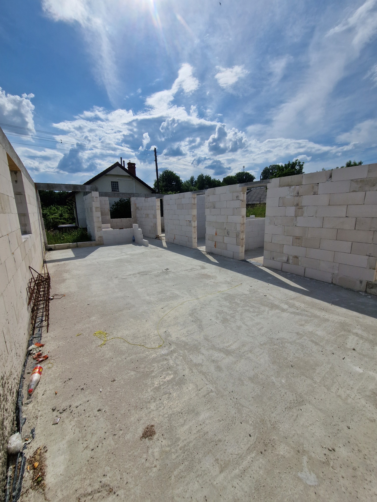 Wnętrze domu w trakcie budowy z białych bloczków, widoczne ściany działowe i betonowa podłoga. W tle dom jednorodzinny i błękitne niebo z chmurami.