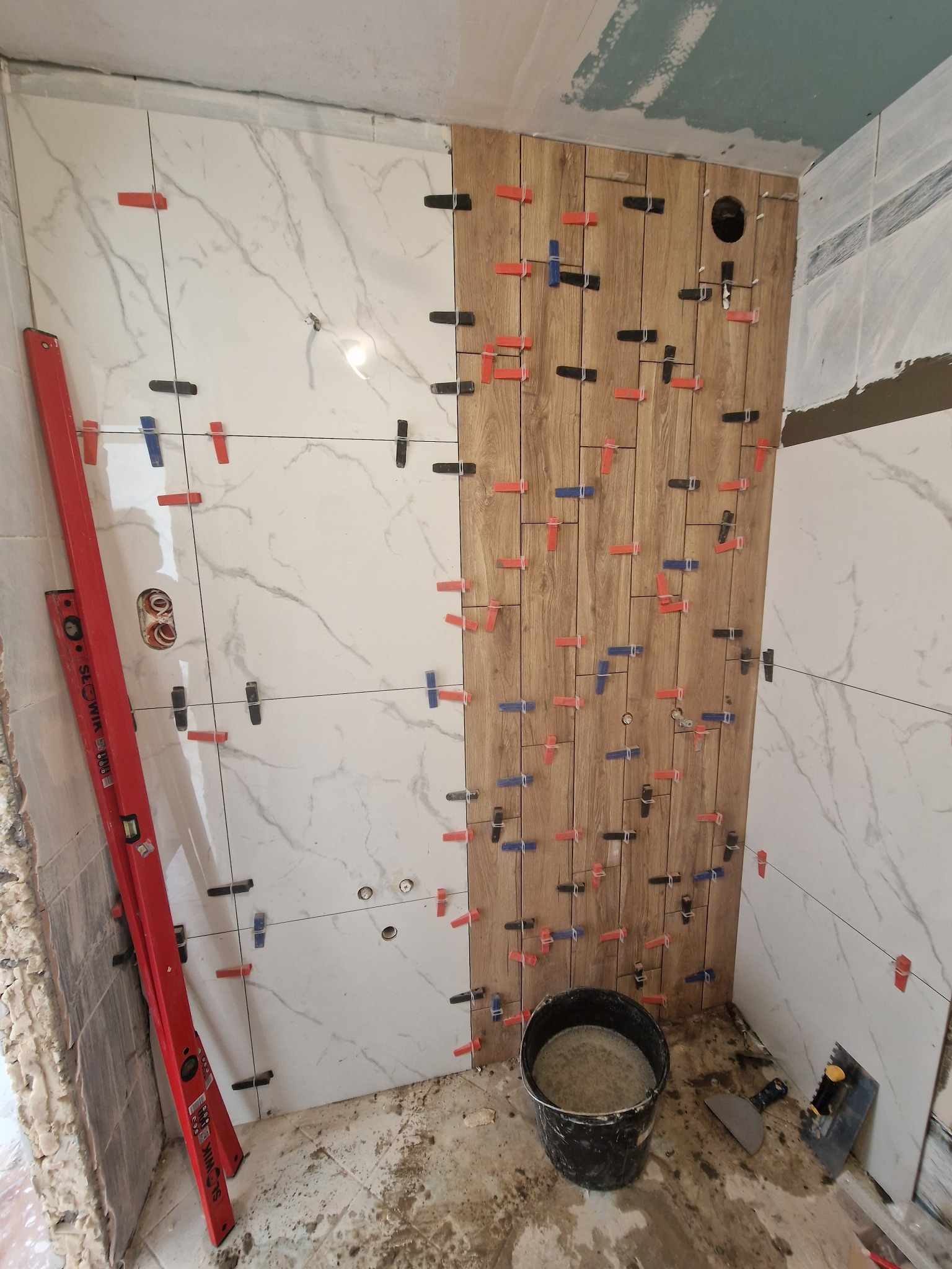 Układanie płytek imitujących drewno i marmur na ścianie z użyciem klipsów poziomujących. Widoczny poziomica i wiadro z zaprawą.