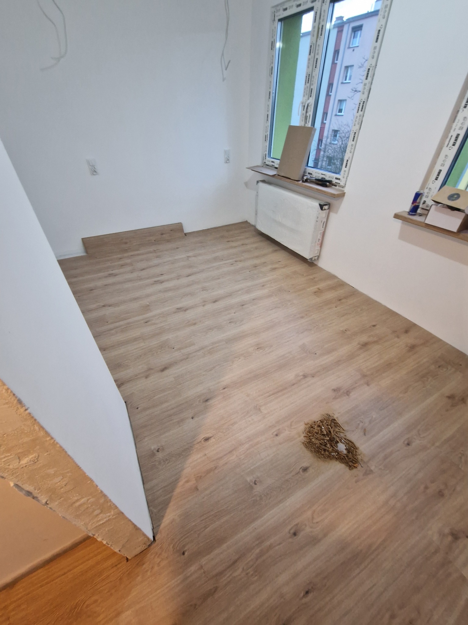 Jasne panele podłogowe ułożone w pokoju z białymi ścianami i oknem. Widoczne resztki po montażu. Fragmenty paneli przy ścianie czekają na montaż.