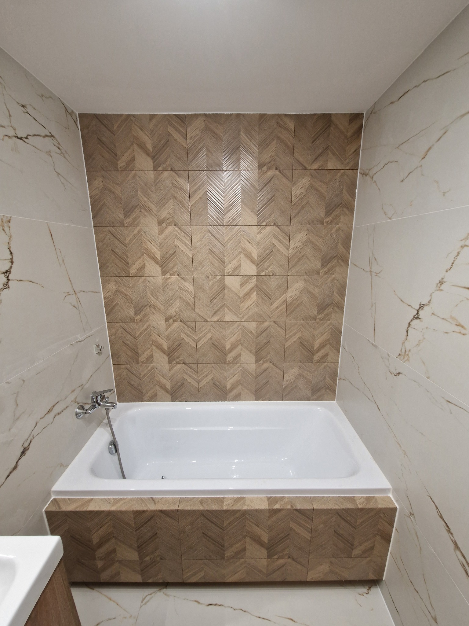 Nowoczesna łazienka z białą wanną obudowaną drewnopodobną mozaiką w jodełkę, otoczoną marmurowymi płytkami z wyraźnym żyłowaniem. Chromowana bateria wannowa.