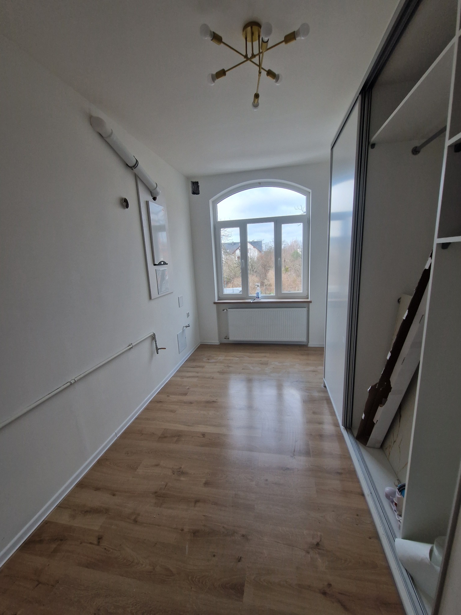 Jasne wnętrze po remoncie w Głazowie: panele podłogowe, białe ściany z instalacjami, okno łukowe i szafa przesuwna. Nowoczesne oświetlenie sufitowe.