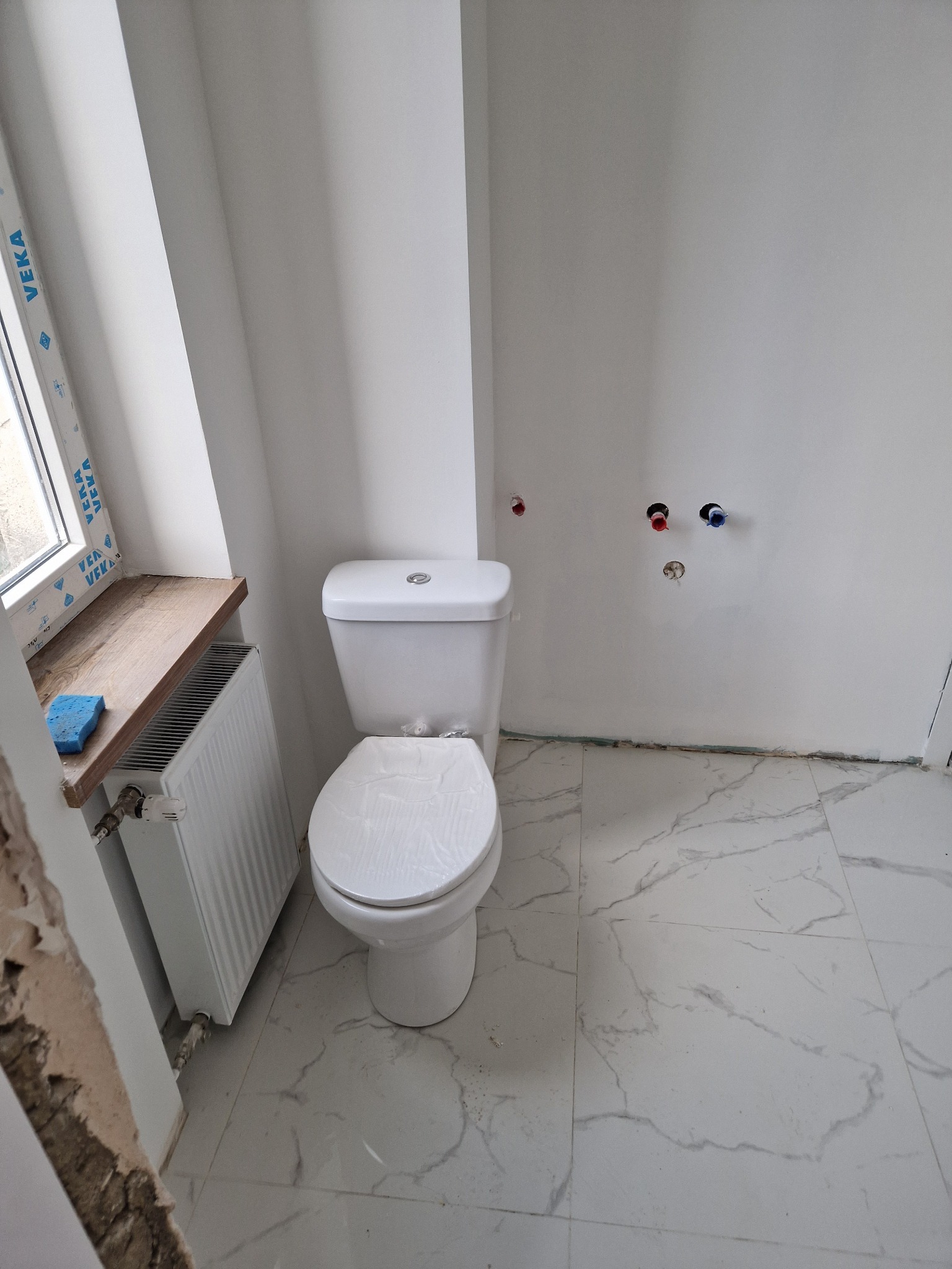 Biała toaleta kompaktowa w wyremontowanej łazience z marmurową podłogą, grzejnikiem pod oknem i przygotowanymi przyłączami wod-kan na ścianie.