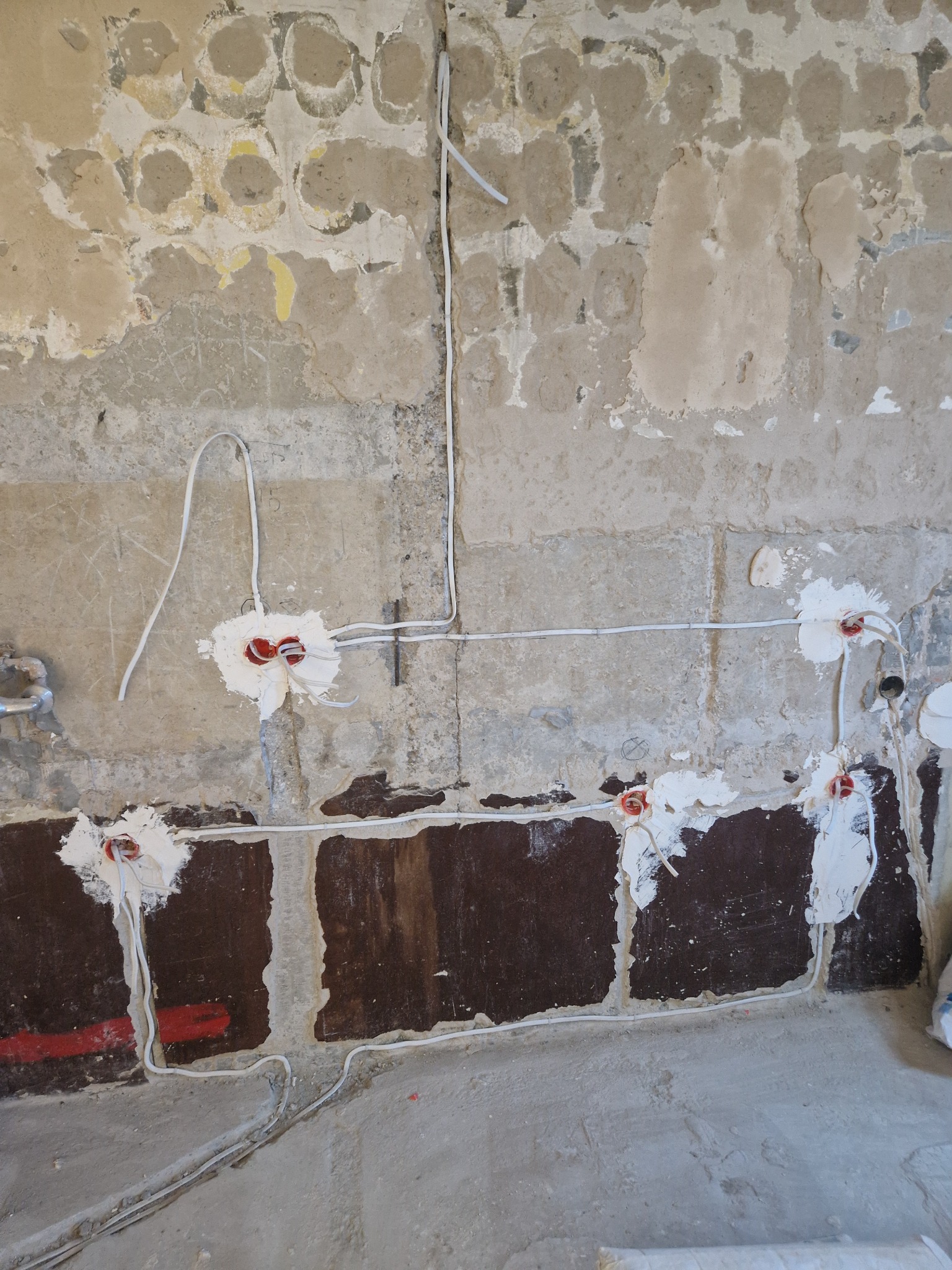 Instalacja elektryczna w trakcie prac: białe kable prowadzone po surowej ścianie z widocznymi puszkami instalacyjnymi, zamaskowanymi gipsem. Widoczne ślady po starej farbie.