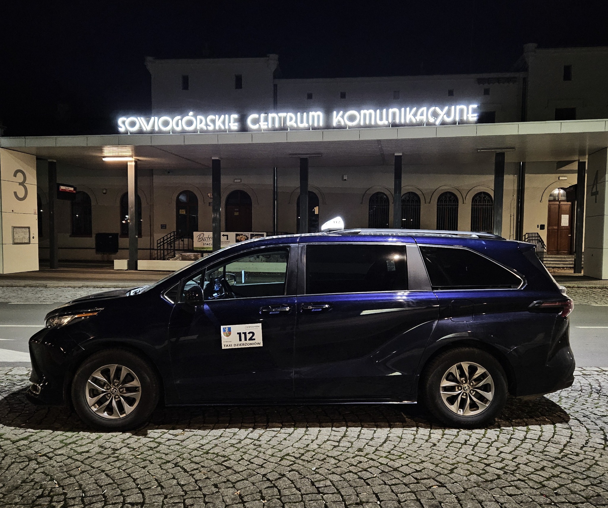 Niebieski van taxi z napisem 'TAXI Dzierżoniów 112' stoi na brukowanej ulicy przed budynkiem 'Sowiogórskie Centrum Komunikacyjne' nocą.