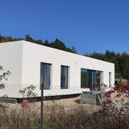 Sokół - Nowoczesny, parterowy dom z białymi ścianami i niebieskimi ramami okiennymi, otoczony zielenią. Widoczne zabezpieczenia okien folią i drabina oparta o ścianę.