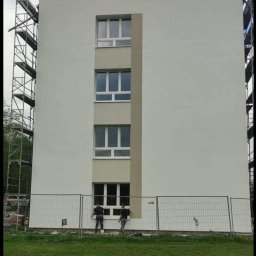 Sokół - Dwóch pracowników maluje elewację budynku z rusztowaniem. Widoczne okna i ogrodzenie. Jasna kolorystyka budynku z szarymi akcentami. Prace wykończeniowe na zewnątrz.