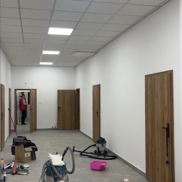 Sokół - Korytarz w trakcie prac wykończeniowych: białe ściany, sufit z kasetonów, drewniane drzwi. Na podłodze narzędzia, drabina i odkurzacz przemysłowy. Widoczna osoba w oddali.