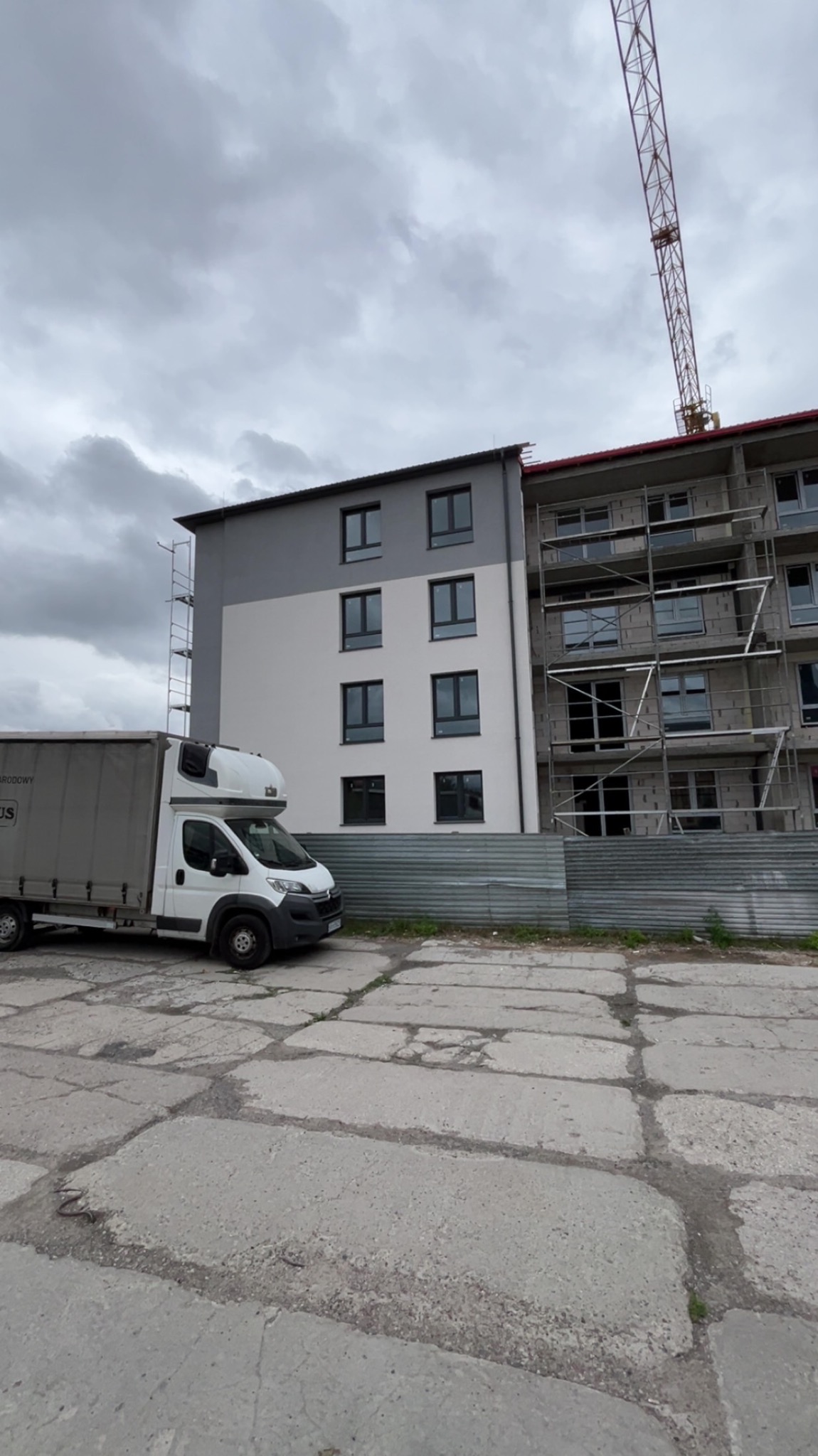 Budynek po częściowym malowaniu elewacji, z widocznym rusztowaniem i dźwigiem w tle, obok zaparkowany biały samochód dostawczy. Szaro-biała kolorystyka budynku.