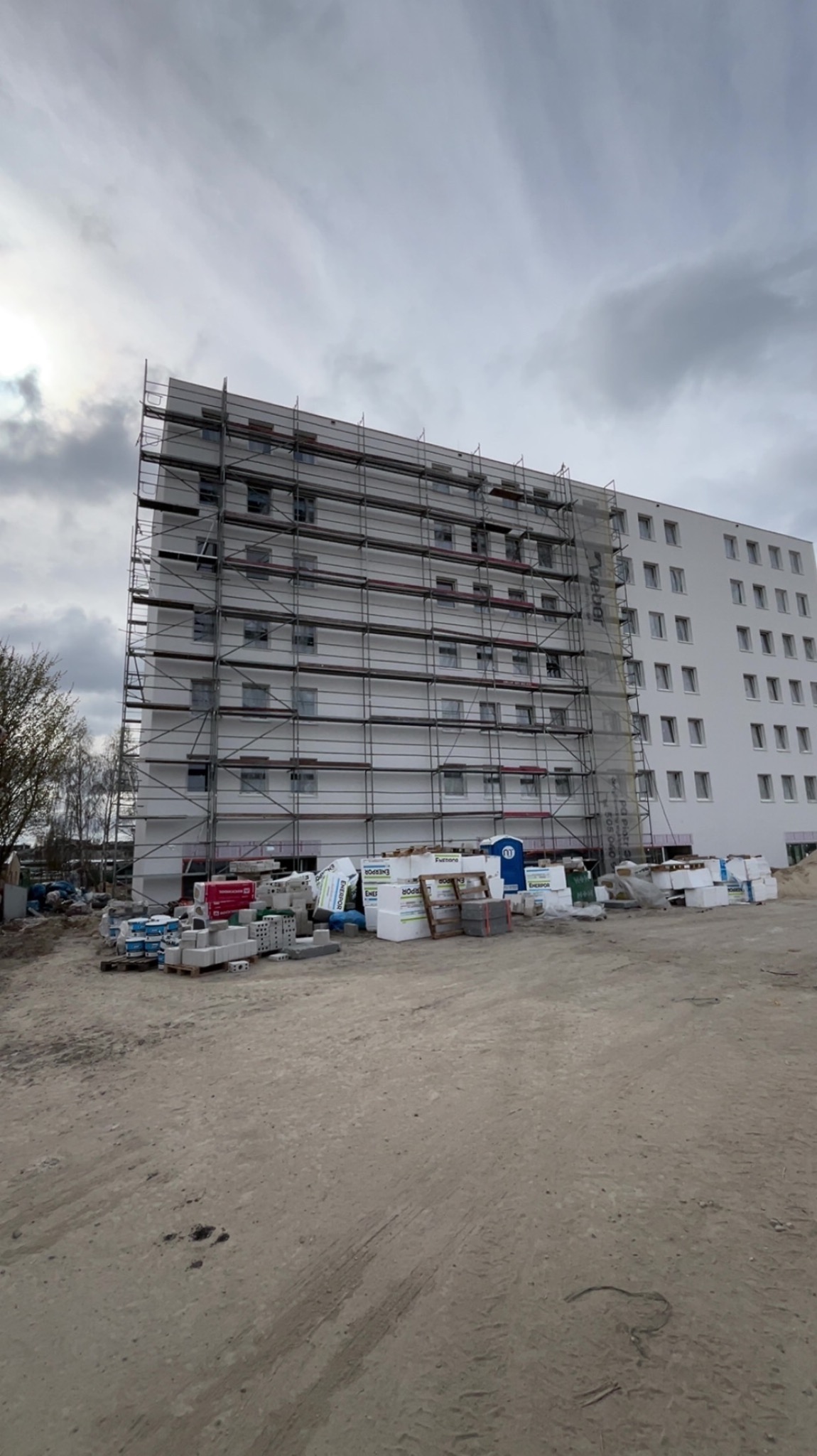 Wielopiętrowy budynek w trakcie prac elewacyjnych, otoczony rusztowaniami i materiałami budowlanymi, na tle pochmurnego nieba. Widoczne okna i świeżo pomalowana fasada.