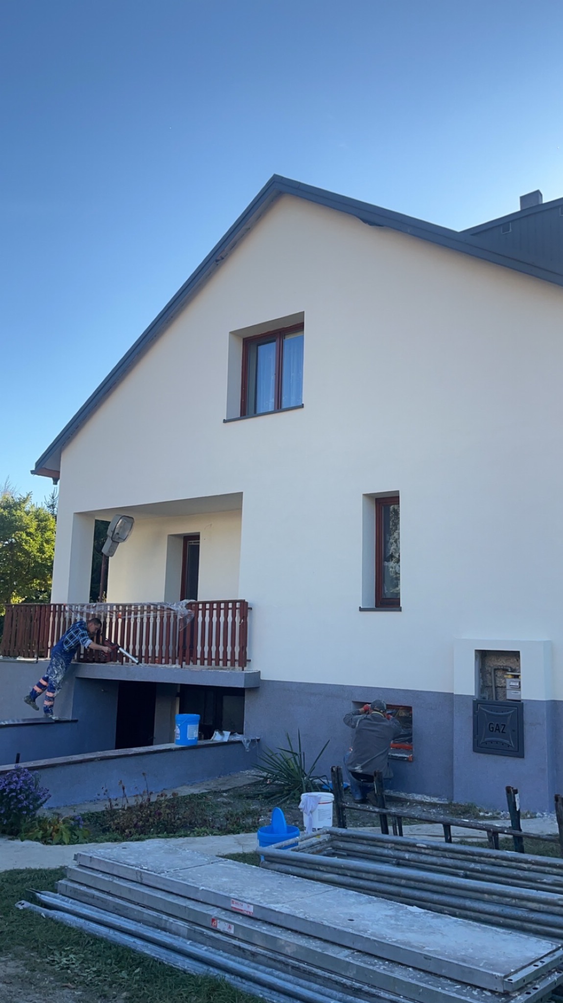 Malowanie elewacji domu jednorodzinnego przez dwóch pracowników. Jeden maluje z balkonu, drugi przyłącze gazowe. Widoczne rusztowania i narzędzia malarskie.
