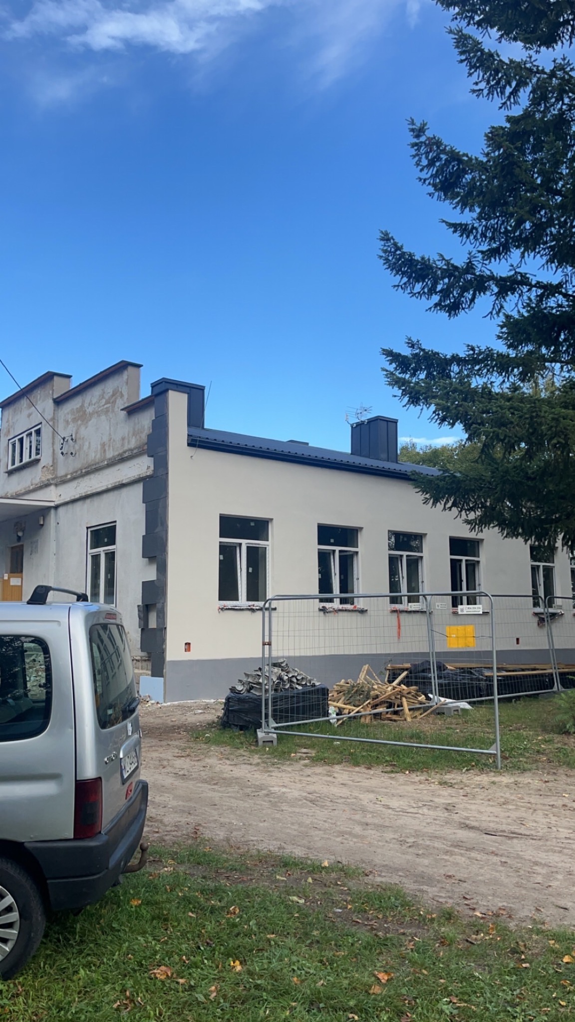 Budynek po częściowej renowacji elewacji, z widocznym fragmentem starej i nowej fasady. Na pierwszym planie zaparkowany samochód i ogrodzenie budowy.