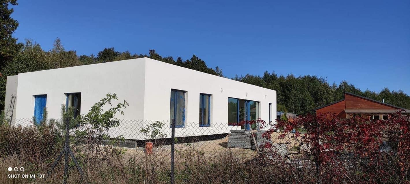 Nowoczesny, parterowy dom z białymi ścianami i niebieskimi ramami okiennymi, otoczony zielenią. Widoczne zabezpieczenia okien folią i drabina oparta o ścianę.