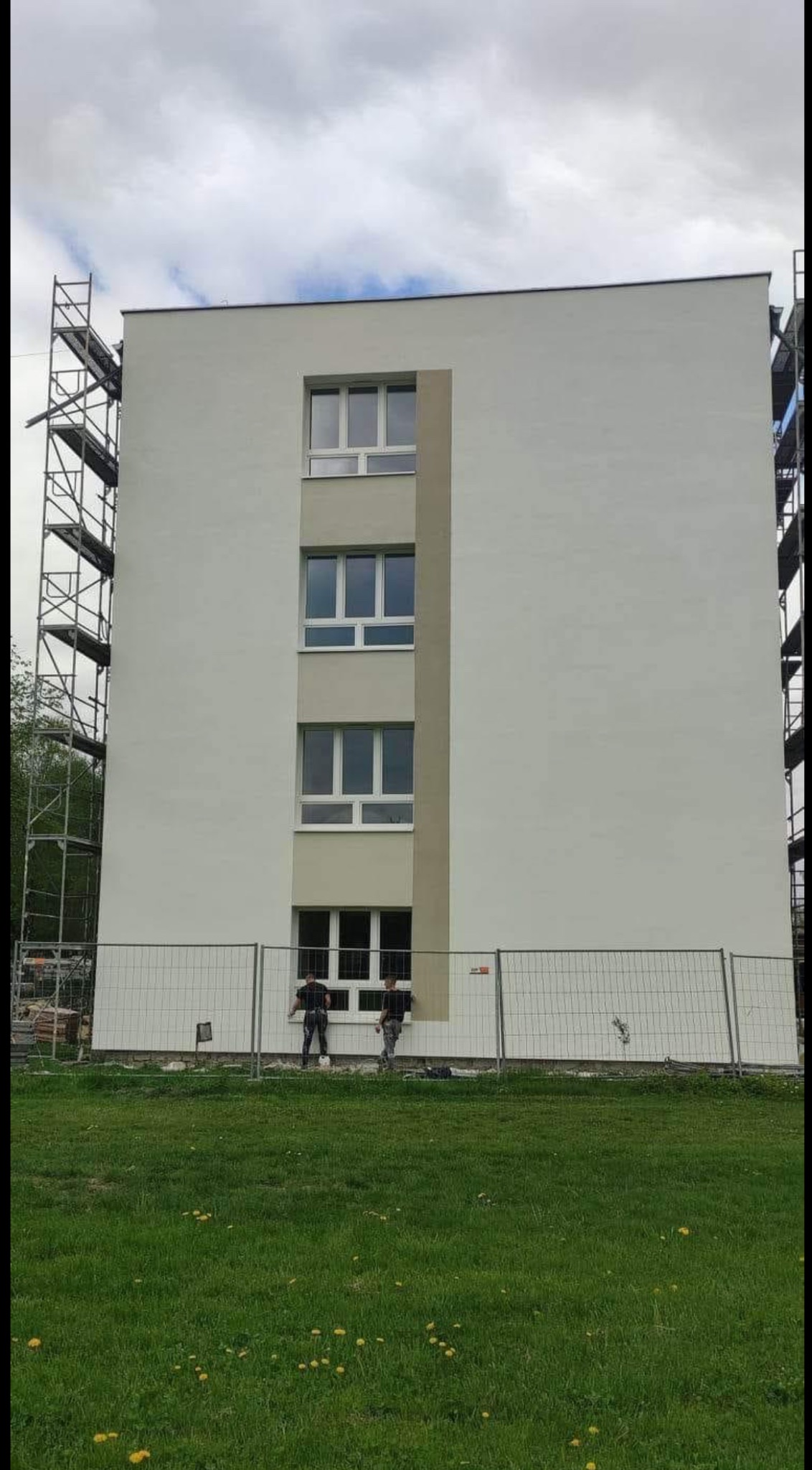 Dwóch pracowników maluje elewację budynku z rusztowaniem. Widoczne okna i ogrodzenie. Jasna kolorystyka budynku z szarymi akcentami. Prace wykończeniowe na zewnątrz.