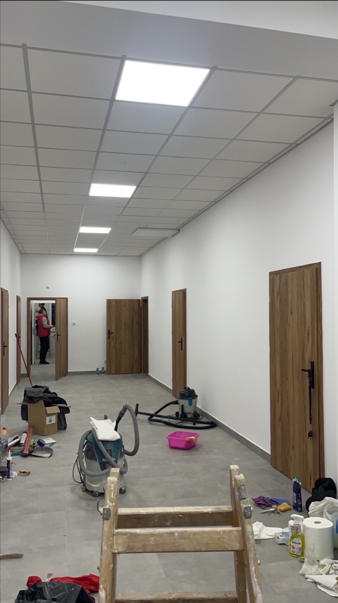 Korytarz w trakcie prac wykończeniowych: białe ściany, sufit z kasetonów, drewniane drzwi. Na podłodze narzędzia, drabina i odkurzacz przemysłowy. Widoczna osoba w oddali.