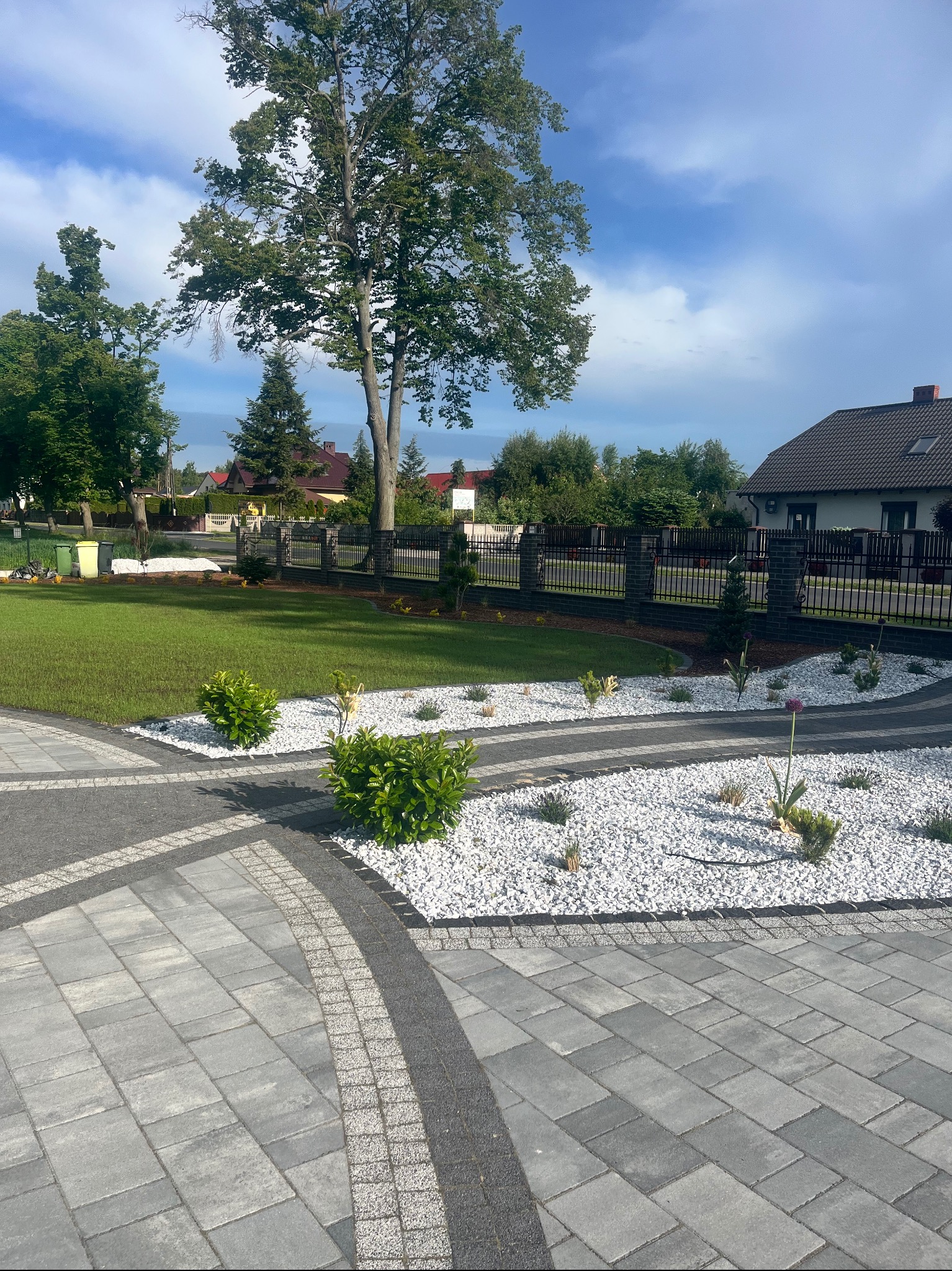 Nowoczesny ogród z szarym brukiem, białym żwirem i zielonym trawnikiem. Widoczne krzewy i drzewa, ogrodzenie z cegły i metalu. Słoneczny dzień, błękitne niebo.