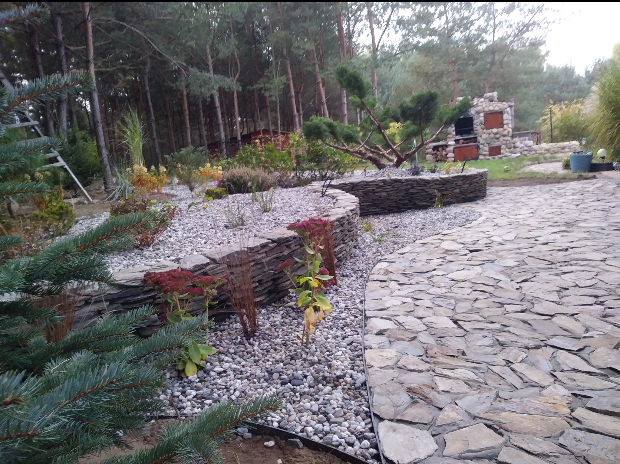 Ogród z kamienną ścieżką i rabatami, otoczony lasem. Kamienne murki oddzielają rabaty wysypane żwirem. W tle kamienny grill.