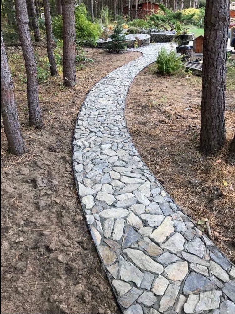 Kręta ścieżka z nieregularnych kamieni w leśnym ogrodzie. Naturalny kamień w odcieniach szarości tworzy harmonijną kompozycję z otaczającą zielenią i drzewami.