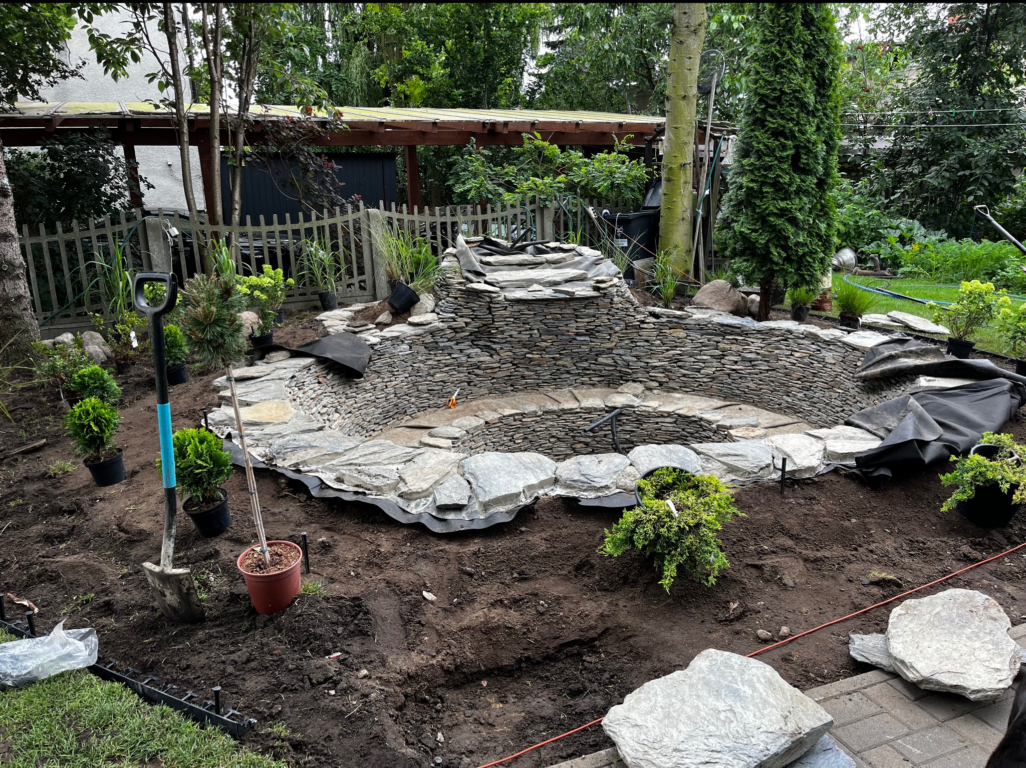 Budowa oczka wodnego z kaskadą w ogrodzie. Widoczne kamienie, rośliny w doniczkach, narzędzia ogrodnicze i folia izolacyjna. Etap prac ziemnych i układania kamieni.