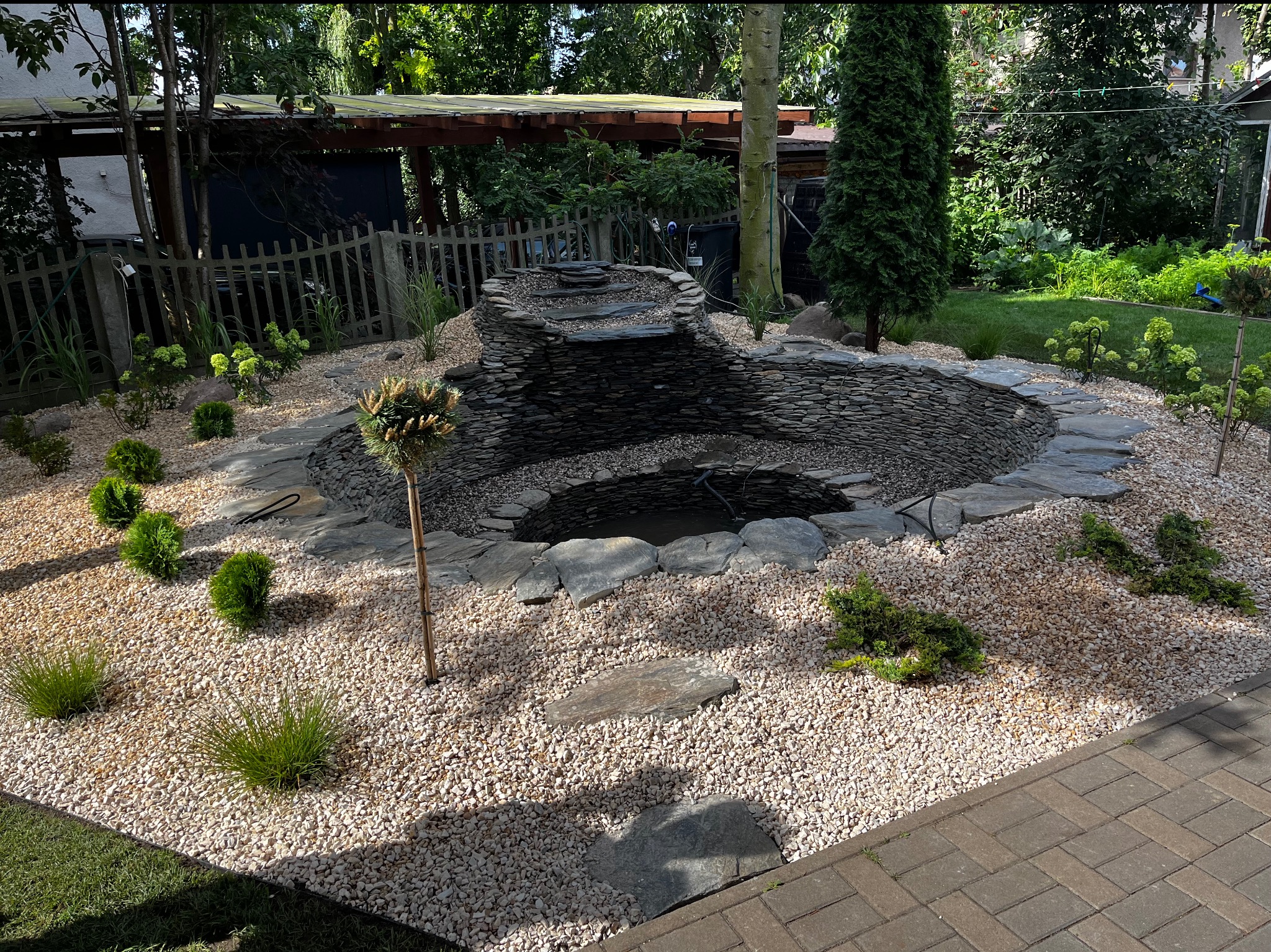 Oczko wodne z kaskadą obłożone kamieniem, otoczone białym żwirem i niską zielenią. Ścieżka z kamiennych płyt prowadzi do oczka. W tle zielony ogród.