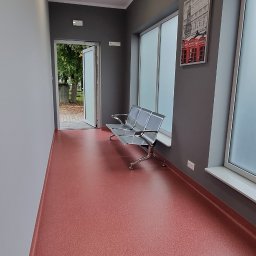 Sebastian Domin - Wnętrze korytarza z czerwoną podłogą, szarymi ścianami, metalową ławką i oknami. Na ścianie wisi obraz z motywem budki telefonicznej.