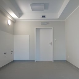 Sebastian Domin - Wnętrze po remoncie: białe ściany, szara podłoga z listwami, białe płytki na ścianie z instalacją wodną, białe drzwi. Minimalistyczny design, neutralna kolorystyka.