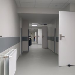 Sebastian Domin - Jasny korytarz w biurze lub placówce medycznej po remoncie, z białymi ścianami, szarymi pasami ochronnymi i grzejnikiem. Widoczne drzwi i oświetlenie sufitowe.