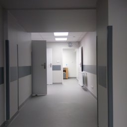 Sebastian Domin - Korytarz w neutralnych barwach z szarymi panelami ściennymi i drzwiami. Widoczny zegar na końcu korytarza, oświetlenie sufitowe i grzejnik.