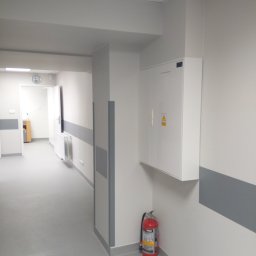 Sebastian Domin - Wnętrze korytarza w odcieniach szarości i bieli z widoczną szafką elektryczną i gaśnicą. Ściany zabezpieczone szarymi pasami. W tle widoczne drzwi i zegar.