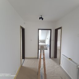 Sebastian Domin - Białe wnętrze korytarza po malowaniu, zabezpieczone folią i taśmą malarską, z rozstawioną drabiną. Widoczne drzwi z ciemnymi futrynami i grzejnik.