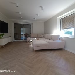 Sebastian Domin - Jasne wnętrze salonu z parkietem w jodełkę, narożną sofą i minimalistyczną szafką RTV. Widoczne okno z roletami i przesuwne drzwi balkonowe.