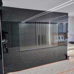 Sebastian Domin - Nowoczesna ściana z czarnego szkła z wbudowanym ekranem i oświetleniem LED. Widoczne elementy instalacji elektrycznej i schody z podświetleniem w trakcie wykończenia.