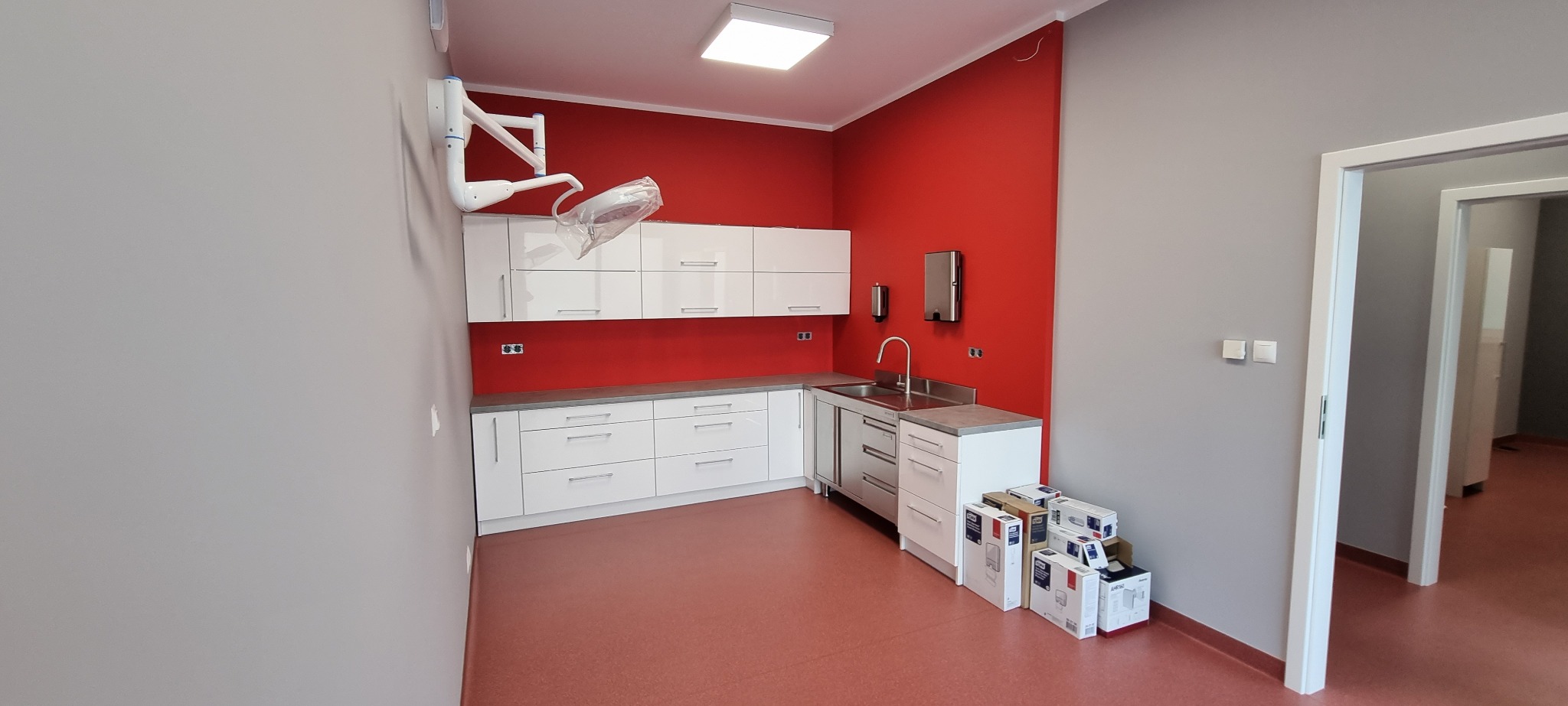Wyposażone wnętrze gabinetu lekarskiego z czerwonym akcentem: białe szafki, umywalka ze stali nierdzewnej, lampa zabiegowa i podłoga w odcieniach czerwieni.