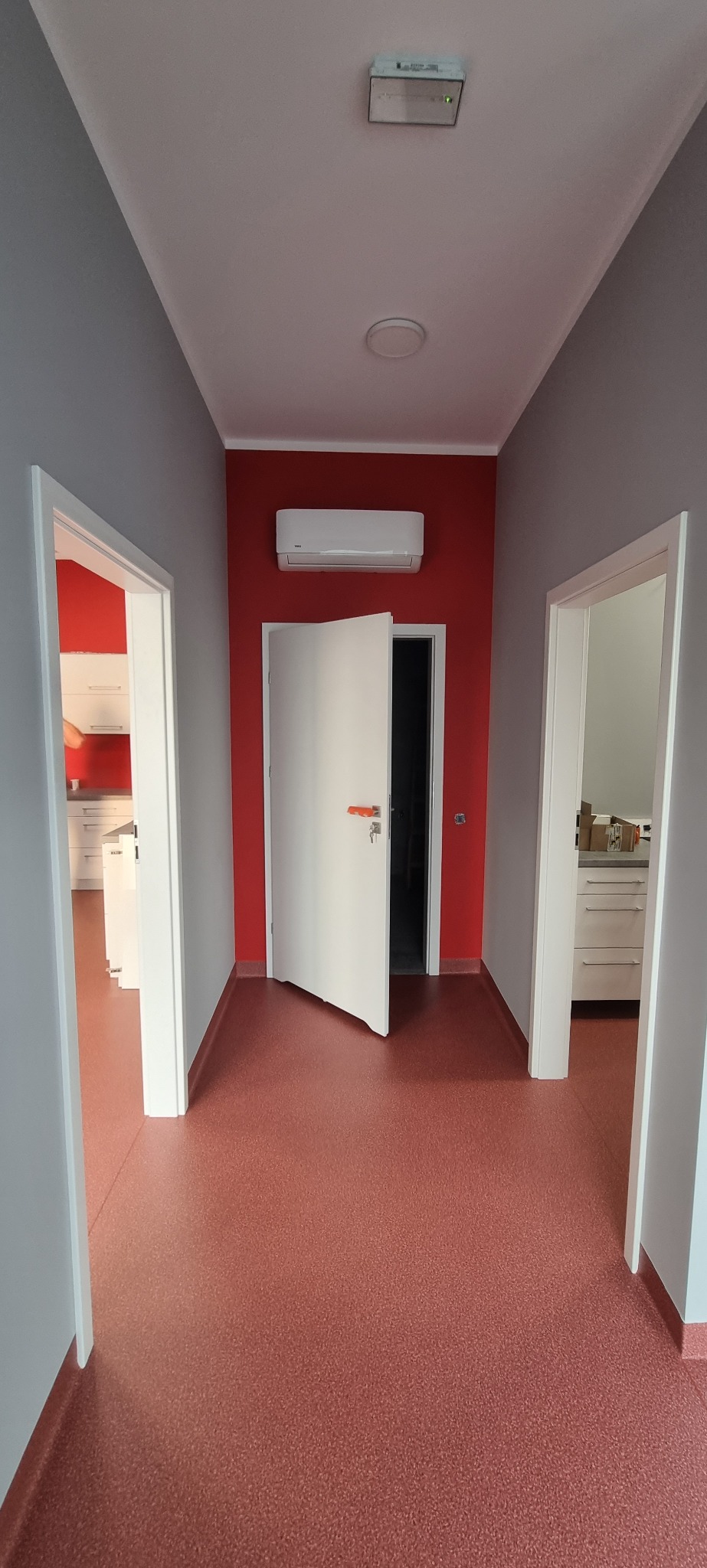 Korytarz po remoncie z czerwonymi ścianami, białymi drzwiami i klimatyzacją. Widoczne wejścia do pomieszczeń z białymi meblami. Podłoga w odcieniach czerwieni.