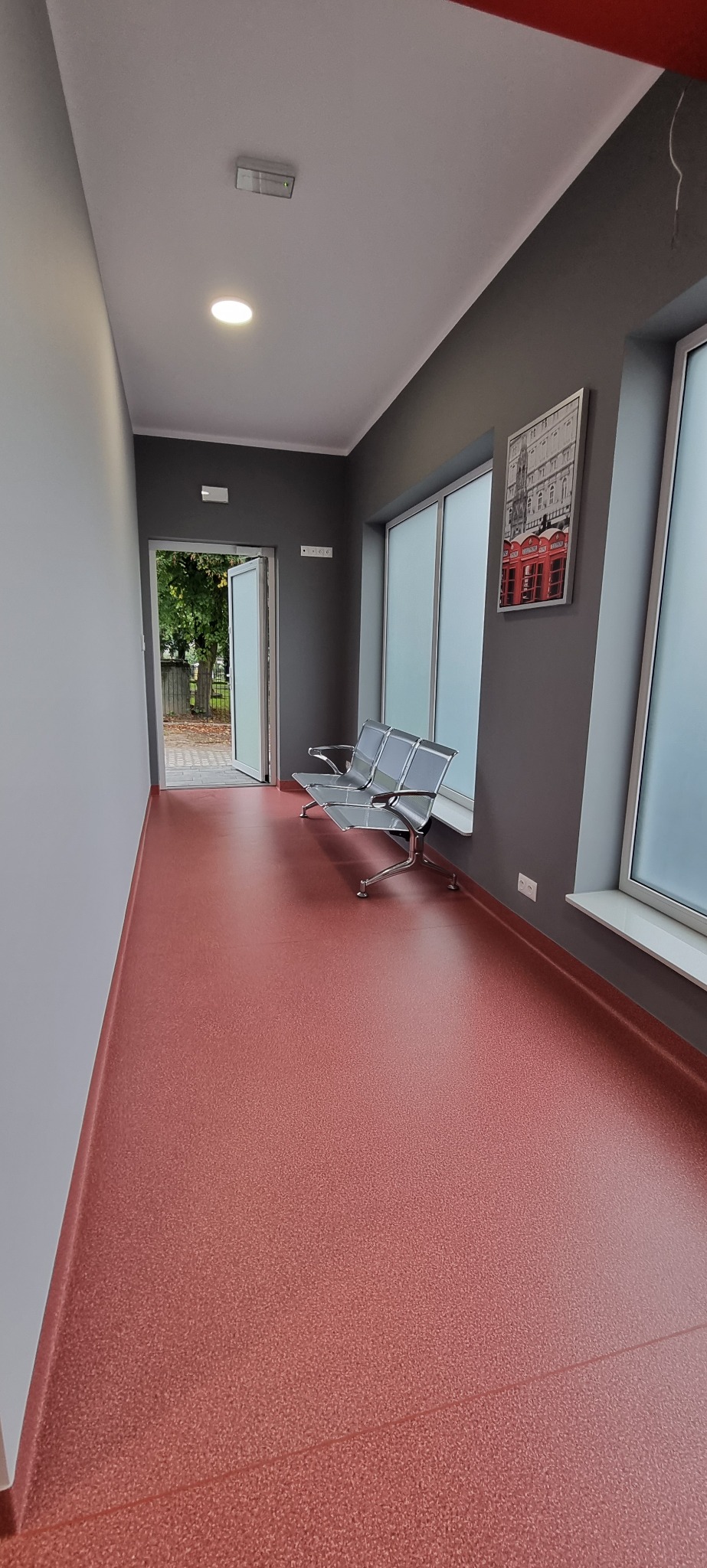 Wnętrze korytarza z czerwoną podłogą, szarymi ścianami, metalową ławką i oknami. Na ścianie wisi obraz z motywem budki telefonicznej.